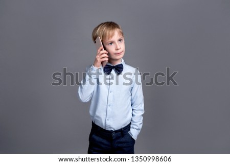 
Little blond boy wearing a blue shirt on a gray background attentively listening to a business phone call holding the phone near the ear, a children’s startup