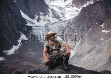 a middle-aged man in a cowboy hat sits on a rock against a glacier and a mountain Altai