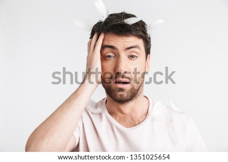 Image of awakened man 30s with bristle in casual t-shirt standing under falling feathers isolated over white background