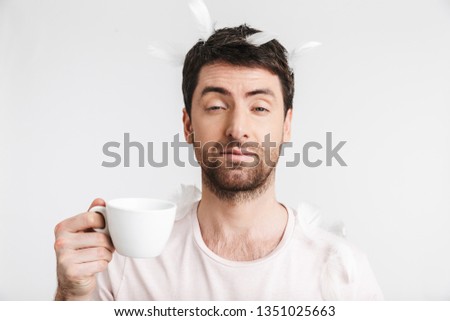 Image of morning man 30s with bristle in casual t-shirt drinking coffee while standing under falling feathers isolated over white background