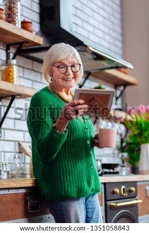 Happy granny with photo frame. Charming stylish beaming granny with short silver hairstyle feasting eyes with grandchildren picture in frame leaning on ready-built kitchen