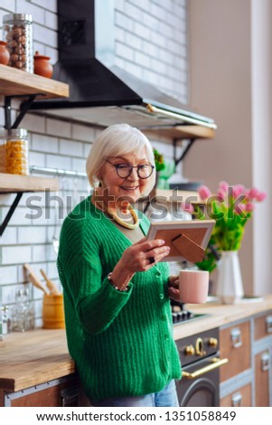 Admiring photo in frame. Graceful silver-haired dame in emerald cardigan and stylish glasses cheerfully admiring photo in frame and holding morning coffee at kitchen