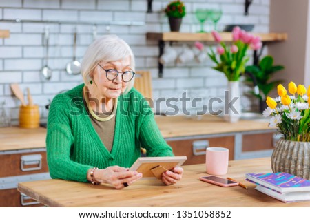 Looking at family portrait in frame. Nice-looking concerned grey-haired dame in knitted green sweater attentively looking at family portrait in frame leaning on wooden kitchen table.