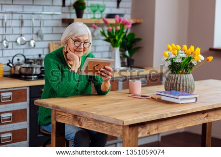 Looking at family photo. Joyful happy aged silver-haired madam wearing bright green-colored apparel delighting in relatives portrait in frame spending time at cozy kitchen