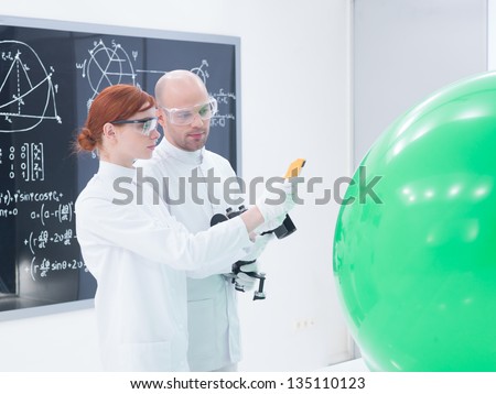 side view of teacher and student in a chemistry lab scanning a green balloon and a  blackboard on the background