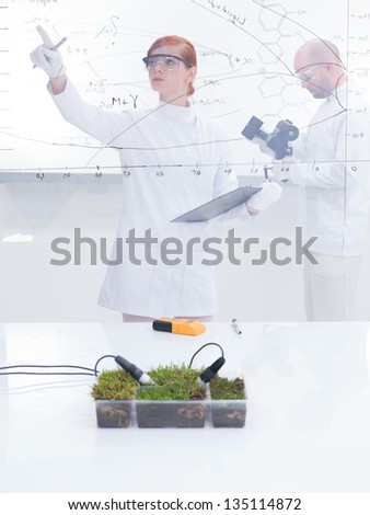 general-view  of a student in a chemistry lab analyzing graphics and her teacher analyzing under microscope in the background of a transparent board around lab table with a box of grass on it
