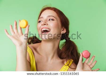Image of a beautiful young redhead girl posing isolated over green wall background holding sweeties macaroons.