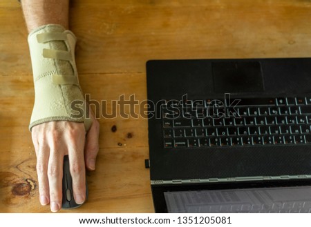 a hand seen wearing a medical hand and wrist brace using a laptop on a wooden table Royalty-Free Stock Photo #1351205081