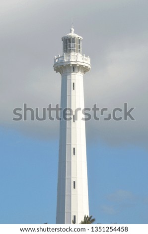 The tower of a white light house rises against a blue sky. Grey clouds fill the top of the picture. Small vertical windows are in the tower.