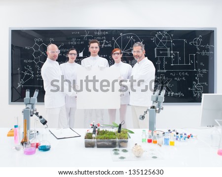 general-view of five confident people in a chemistry lab holding in hands an empty banner around lab tools , leafs and colorful liquids with a blackboard with formulas  on the background