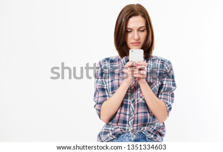 Closeup photo of casually-dressed European girl standing isolated on white background looking attentively at screen of cellphone, browsing web pages and smiling nicely while chatting with friends.