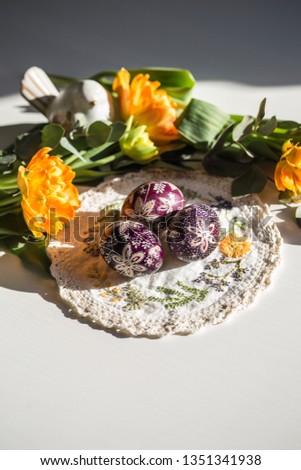 Three Easter painted eggs on handmade embroidered napkin with orange tulips and eucalyptus branches on background on white wooden table.
