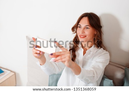 Young caucasian smiling pretty woman with curly brown hair in white shirt taking pictures, selfie with mobile phone, sitting on bed in bedroom, room or apartment, indoors. Horizontal copy space
