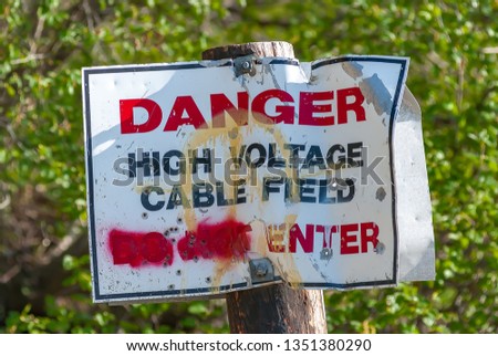 Danger High Voltage Cable Field Do Not Enter Sign in rough shape after being graffitied and shot full of holes.