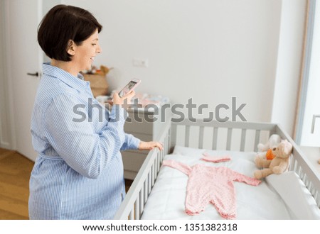 motherhood, pregnancy and nursery concept - happy pregnant middle-aged woman with smartphone ptotographing baby clothes on bed at home