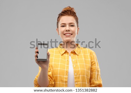 technology, communication and people concept - smiling red haired teenage girl in checkered shirt showing blank screen of smartphone over grey background