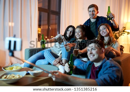 friendship, people, technology and party concept - happy friends with non-alcoholic drinks taking picture by selfie stick at home in evening