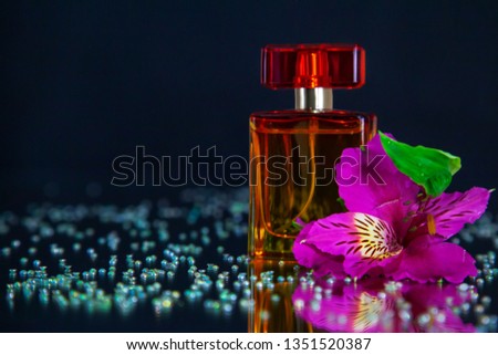 Macro photography of a bottle of perfume standing on a mirror near a pink flower among rhinestones. Studio photography close up on a black background, using turquoise backlighting.