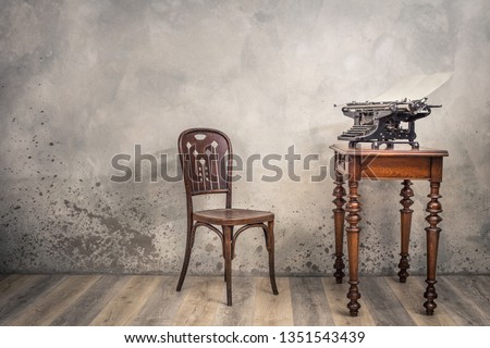 Vintage loft room with old typewriter on wooden table and antique chair front concrete wall background with shadows. Retro style filtered photo