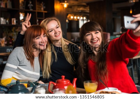 Three girlfriends make selfie in a restaurant, in a diner, sisters, blonde, brunette and red, tea and desserts, happy, fooling around