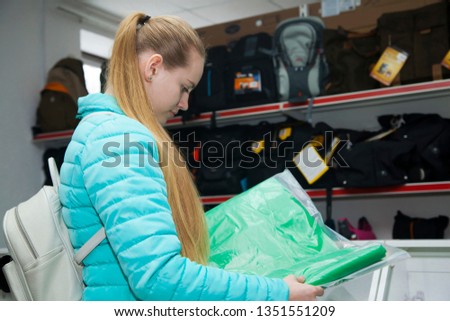 Young blonde in professional photo equipment store chooses the green fabric for background of studio. Standing in profile.