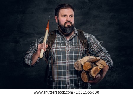 Portrait of a bearded woodcutter with a backpack dressed in a plaid shirt holding firewood and ax. Studio photo against a dark textured wall