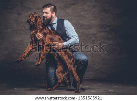 Middle-aged hunter dressed in elegant clothes sits on his knee and holding his purebred brown Setter. Studio photo against a dark textured wall