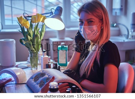 Master cosmetologist in mask and gloves sits at the manicure table and looks at the camera. Photo with dim light and red Blue backlight