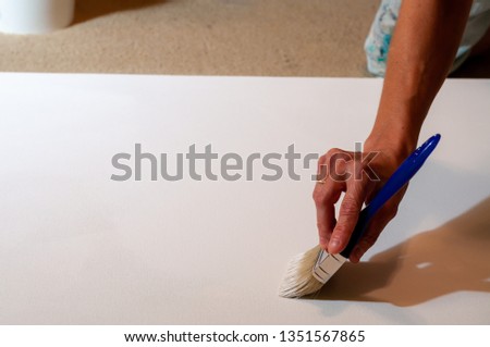 Female artist's hand applying white primer coating on a canvas on her studio floor.
