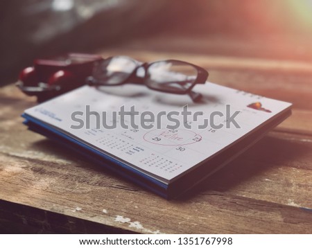 White paper calendar has note stuck on an calendar, red Alarm clock,and eyeglasses on empty wood planks.Written reminder to Retire.Elderly memories concept,with dark tone,dimly light.Selective focus.