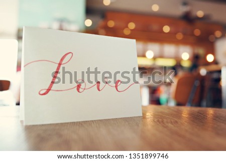 Valentine card with "LOVE" hand lettering display on wooden desk, blurred cafe interior background.                