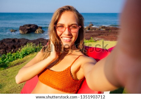 happy smiling girl in a stylish orange coral swimsuit taking selfie on phone in vacation. bikini woman holding cellphone photo camera smartphone pictures app sitting on a lounger on the ocean india
