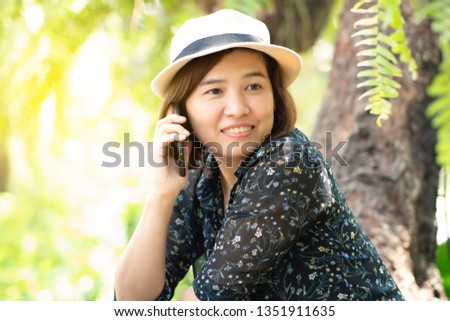 Asian woman wearing a hat sitting in the park, she is talking on the phone, she smiles happily. Fresh air makes her feel good.