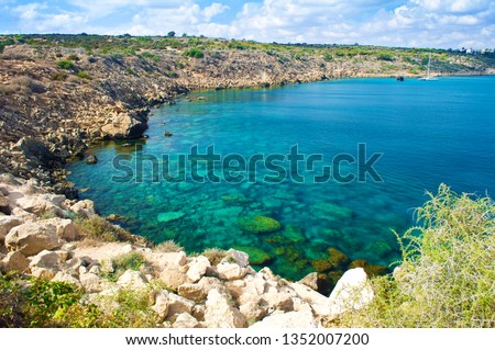 Image with a view of a bay near Cape Greco, Cyprus. Rock coastline near deep green transparent emerald water against a rocky hill. Amazing cloudscape. Warm day in fall