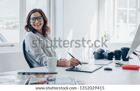 Professional photographer sitting at her office desk looking away and smiling. Woman in office with digital graphic tablet and drawing pen.