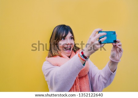A brunette woman is in the street next to a yellow wall and a photo is taken with her mobile. Look at the camera with surprise and happiness while holding the phone with both hands. 
