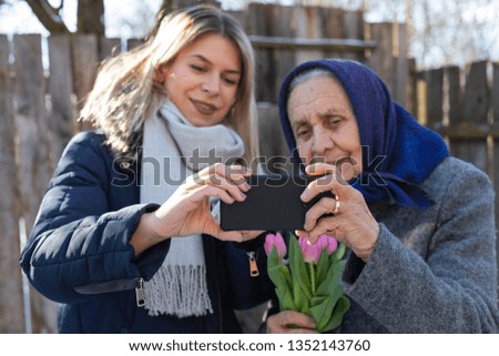 Picture of joyful women taking a selfie outdoor - young beautiful woman with old granny holding a smart phone and flower bouquet, smiling to the camera