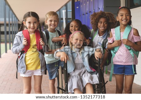 Front view of happy diverse school kids standing in  outside corridor at school while a Caucasian schoolgirl is sitting on wheelchair in foreground Royalty-Free Stock Photo #1352177318