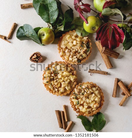 Homemade sweet apple shortbread tartlets with cinnamon sticks, walnuts, apples, red leaves branches above over white marble background. Flat lay, space. Square image