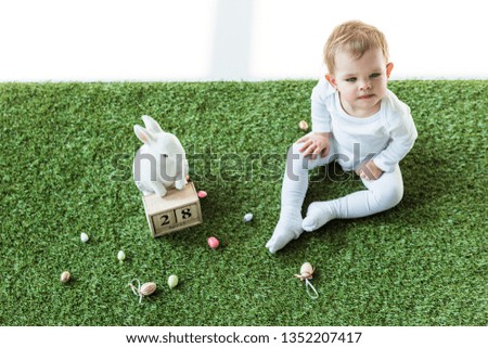 cute baby sitting on green grass near wooden calendar with 28 April date, decorative rabbit and colorful quail eggs isolated on white