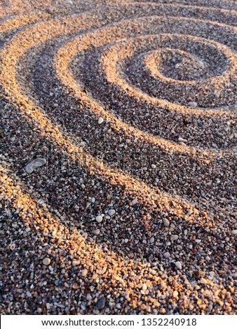 Spiral line drawn in rough sea sand. Small rocks on sea shore. Sand texture reflecting the evening sun.