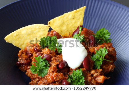 Chili con carne meal in a rustic bowl. Traditional dish of mexican cuisine with kidney beans, minced meat, parsley, tortilla chips and sour cream. Close up stock photo.