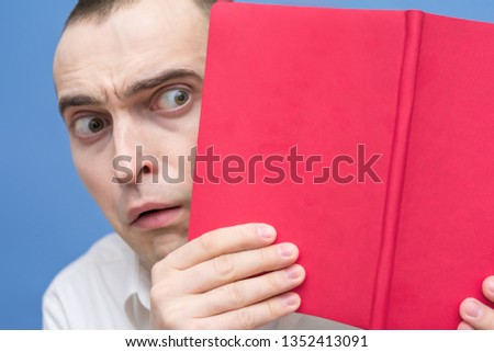 Man looks out from behind an open book, male scared, cover of the red book, close up, background, copy space, for advertising, slogan