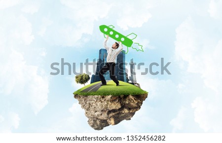 Conceptual image of young confident businessman in suit launching big rocket from his hand while standing on the flying island with cloudy skyscape view on background.