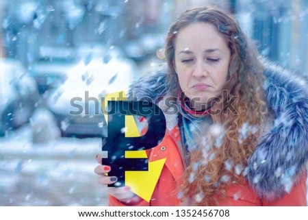 Exaggeratedly sad red-haired girl in warm clothes in winter with a ruble sign and an arrow down. Concept - over the winter, the ruble fell
