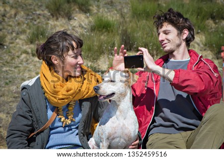 A boy photographs his girlfriend and their dog using a smartphone.