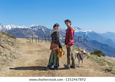 An engaged couple walking on a path in the Italian Alps with their dog on a sunny day.