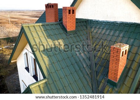 Green metal shingled house roof with attic plastic window and brick chimney.