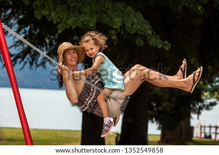 Happy loving family! Young mother and her child daughter swinging on the swings and laughing a summer evening outdoors, beautiful girl on the swing in the park by lake, joyful holidays