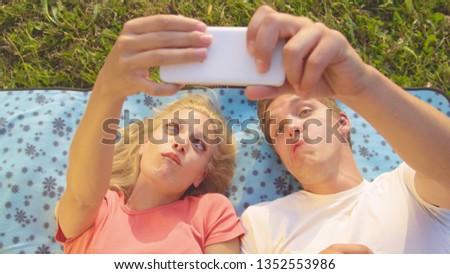 CLOSE UP: Excited blonde haired woman and handsome man making funny faces and taking selfies while lying on a blanket in the middle of a meadow on a warm summer day. Goofy young couple taking photos.